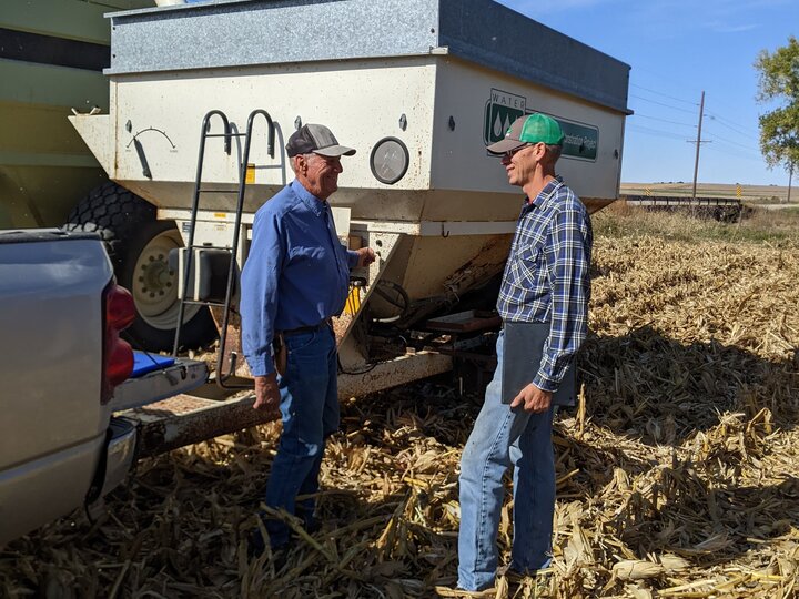 Two farmers in a field standing next to a grain cart